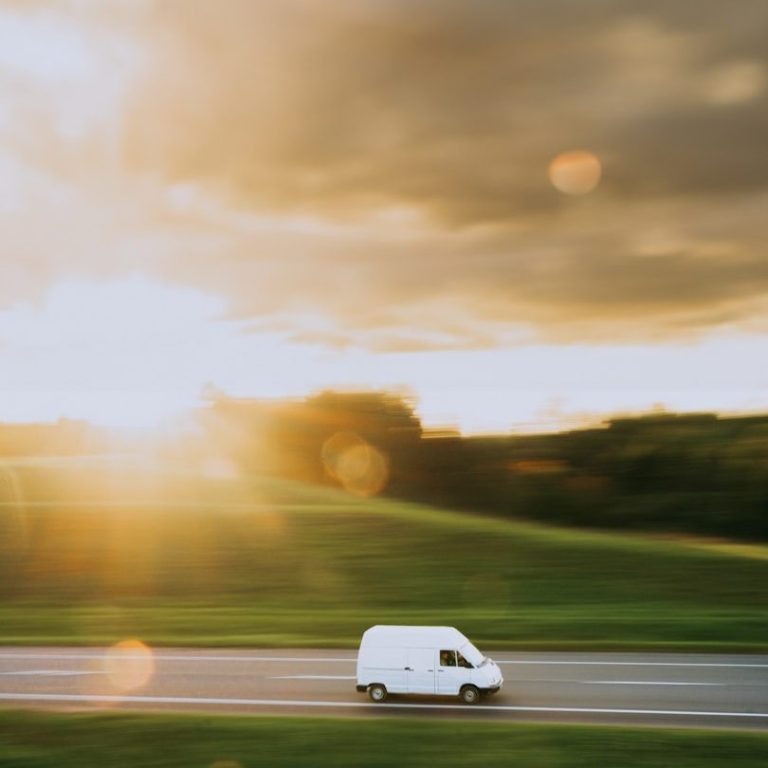 A white van driving on a road at sunset, with golden light and green fields.